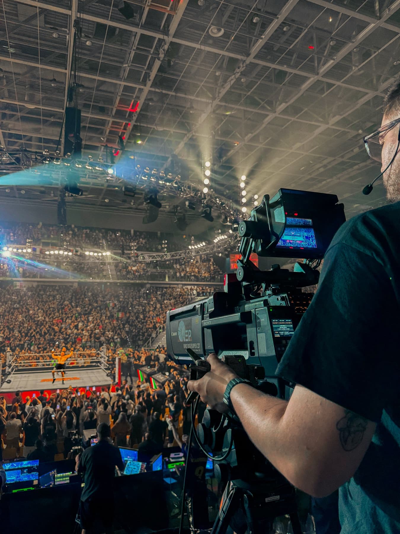 Steve McKinnon operating a broadcast camera with a headset on, the live feed visible on the monitor and the crowd below